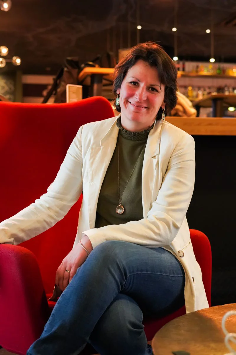 Femme assise sur un fauteuil rouge, souriante, portant une veste blanche et un pull vert.