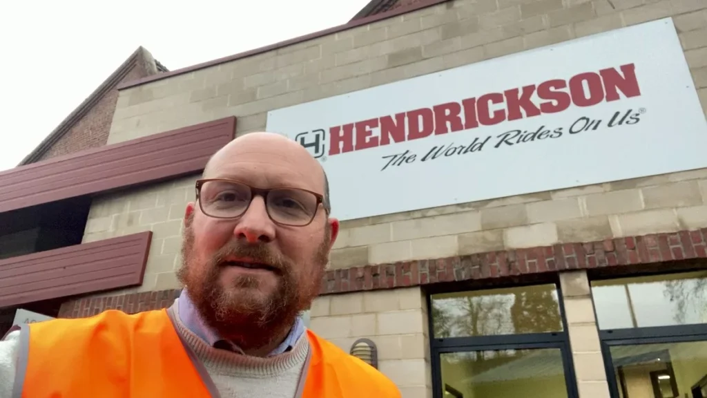 Un homme en gilet orange devant un bâtiment avec un panneau Hendrickson